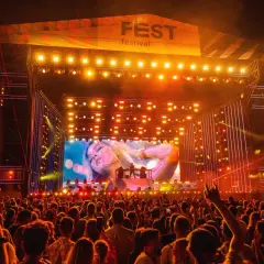 Tłum na festiwalu, scena z wielkim ekranem i logo "FEST festival program". Światła i artyści na scenie.