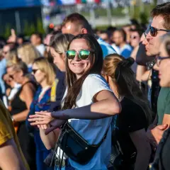 Uśmiechnięta kobieta w okularach przeciwsłonecznych na tle tłumu ludzi na koncercie.
