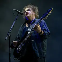 Robert Smith z The Cure na koncercie, w charakterystycznym makijażu, gra na czarnej gitarze elektrycznej.
