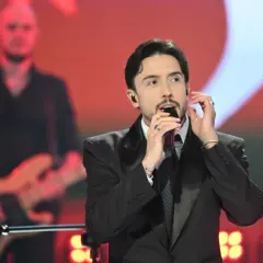 Dawid Kwiatkowski w czarnym garniturze śpiewa do mikrofonu na scenie. W tle muzyk z gitarą.