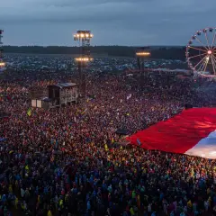 Ogromny tłum ludzi na festiwalu rockowym, rozłożona flaga Polski, scena i diabelski młyn.