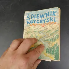 Dłoń trzyma stary śpiewnik harcerski z rysunkiem rzeki i gór.