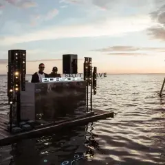 DJ na tratwie podczas zachodu słońca, z napisem "SALT WAVE" i huśtawką na wodzie.