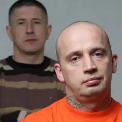 Dwóch mężczyzn, jeden z przodu w pomarańczowej koszulce i tatuażem na szyi, drugi z tyłu w bluzie w paski.