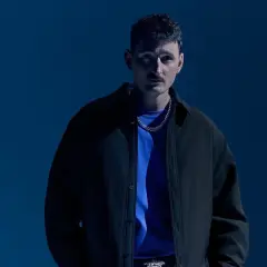 Dawid Podsiadło w czarnej kurtce i niebieskiej koszulce, na tle niebieskiego tła.