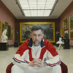 Mężczyzna w dresie siedzi w galerii sztuki, otoczony obrazami i rzeźbami.