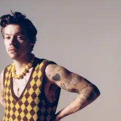 Harry Styles w kamizelce w romby i żółtym naszyjniku, z tatuażami na ramieniu.
