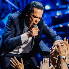 Nick Cave w garniturze śpiewa do mikrofonu, otoczony przez wyciągnięte ręce fanów.