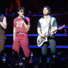 Trzech członków zespołu Jonas Brothers na scenie, jeden śpiewa, drugi gra na gitarze.