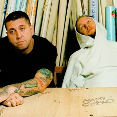 Dwóch mężczyzn siedzi przy stole, jeden z tatuażami na rękach, drugi w białej bluzie z kapturem.
