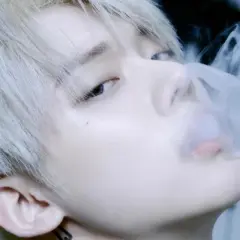 Młody mężczyzna z jasnymi włosami, jeden z członków popularnych k-pop zespołów męskich, wydmuchuje dym.