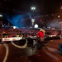 Koncert na scenie z klawiszowcem, tłum ludzi na placu, kamery, światła i dym.