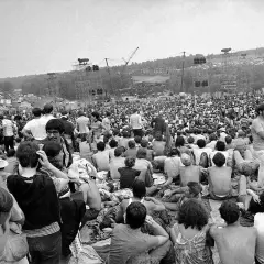 Tysiące ludzi siedzi na trawie, patrząc na scenę festiwalu. To właśnie Woodstock, gdzie jest to legendarne wydarzenie.