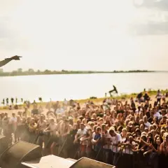 Artysta na scenie podczas jednego z letnich festiwali w Polsce, wskazujący na tłum zgromadzonych fanów nad jeziorem.