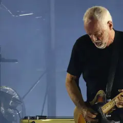 David Gilmour gra na gitarze elektrycznej na tle sceny z dymem i sprzętem muzycznym.