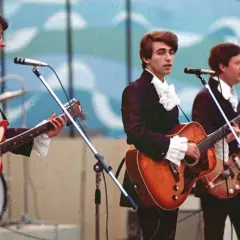 Trzech mężczyzn w garniturach i białych koszulach z żabotami gra na gitarach na scenie.