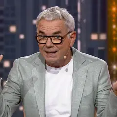 Mężczyzna w okularach i jasnej marynarce gestykuluje, jakby śpiewał z playbacku.