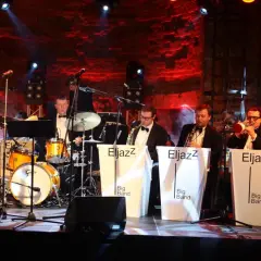 Zespół jazzowy gra na scenie, przygotowując się do sylwestrowych koncertów. Muzycy w eleganckich strojach, instrumenty i czerwone światła tworzą wyjątkową atmosferę.