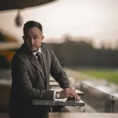 Mężczyzna w garniturze z drinkiem opiera się o barierkę na tle zielonego boiska.
