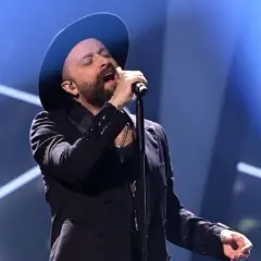 Łukasz Drapała w czarnym kapeluszu i marynarce śpiewa do mikrofonu na scenie.