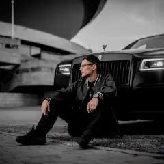 Mężczyzna w czapce i kurtce siedzi obok czarnego Rolls-Royce'a na tle betonowej budowli.