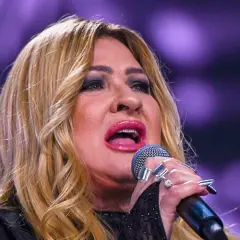 Kobieta z blond włosami śpiewa do mikrofonu, ma jej twarz jest częściowo zasłonięta.