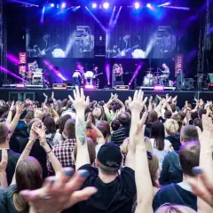 Koncert na żywo, tłum ludzi z podniesionymi rękami, scena z zespołem w tle, fioletowe światła.