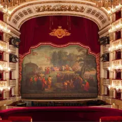 Wnętrze luksusowego teatru operowego z czerwonymi lożami, złotymi zdobieniami i malowidłem na scenie.