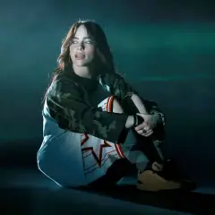 Billie Eilish siedzi na scenie w stroju sportowym, wpatrując się w bok.