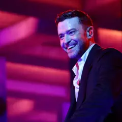Justin Timberlake w czarnym garniturze i białej koszuli, uśmiecha się na tle fioletowo-różowego oświetlenia scenicznego.
