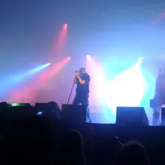 Na scenie podczas koncertu Sisters of Mercy, w dymie i kolorowych światłach, artyści grają na gitarach i śpiewają.