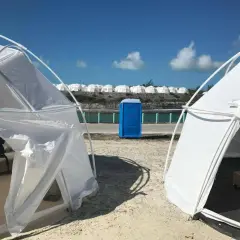 Namioty i niebieska toaleta polowa na plaży, przypominające fiasko Fyre Festival 2. W tle rzędy białych kopuł.