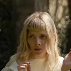 AURORA z blond włosami i grzywką, w białej sukience, na tle zieleni.