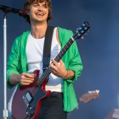 Joe Keery w zielonej koszuli i białej podkoszulce gra na czerwonej gitarze elektrycznej na koncercie.