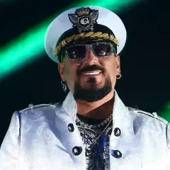 Gigi D'Agostino w białej marynarce i czapce, w okularach przeciwsłonecznych, na tle zielonych świateł.