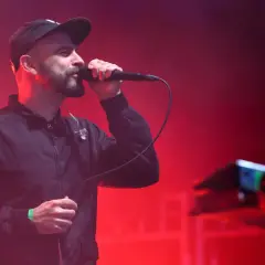 Wokalista z brodą w czapce i kurtce śpiewa do mikrofonu na tle czerwonych świateł. To część tauron nowa muzyka line-up.