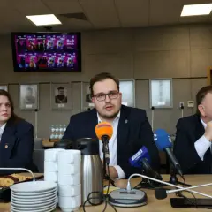 Trzy osoby siedzą przy stole konferencyjnym z mikrofonami i kubkami. Na ekranie w tle widnieje plakat.
