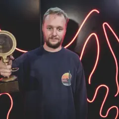 Mężczyzna w granatowej bluzie trzyma złotą statuetkę na tle czerwonych neonów.