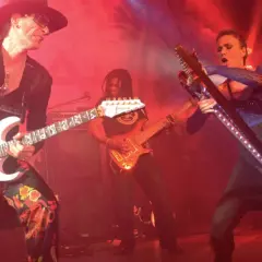 Trzech gitarzystów na scenie, jeden w kapeluszu gra na białej gitarze, drugi w tle na pomarańczowej, trzeci na futurystycznej.