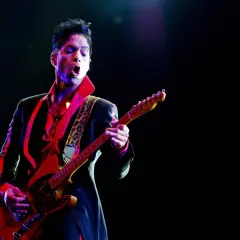 Prince, jako prince opener, gra na gitarze w czerwonej koszuli i czarnej marynarce, oświetlony kolorowymi światłami sceny.