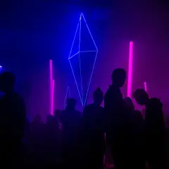 Sylwetki ludzi na tle niebiesko-fioletowych świateł i geometrycznej instalacji świetlnej.