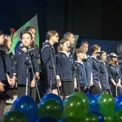 Dzieci w mundurkach śpiewają na scenie, przed nimi mikrofony i balony.