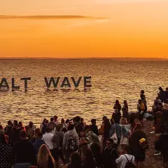 Ludzie na plaży podczas zachodu słońca, z napisem "SALT WAVE" na wodzie.