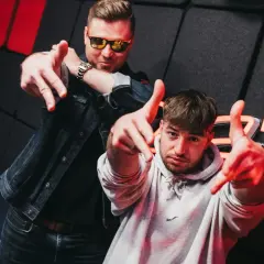 Dwóch mężczyzn pozuje na tle studia radiowego z logo "Zet Radio", wskazując palcami w stronę widza.