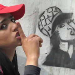 Kobieta w czerwonej czapce z palcem na ustach obok graffiti przedstawiającego kobietę w czapce.
