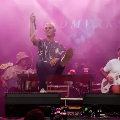 Zespół grający na scenie festiwalowej w różowym świetle. Wokalista siedzi na ławce, trzymając mikrofon.