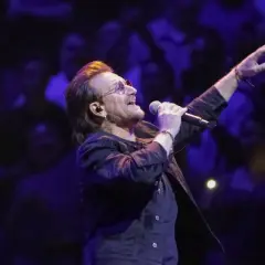 Bono z U2 śpiewa na koncercie, zapowiadając u2 nowa płyta. W tłumie widać światła i sylwetki fanów.