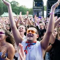 Tłum na koncercie cieszy się letnie brzmienia. Ludzie klaszczą, podnoszą ręce, a niektórzy mają kolorowe okulary i chusty.