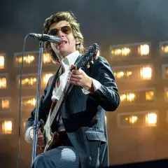 Alex Turner z Arctic Monkeys na scenie, w okularach i garniturze, gra na gitarze podczas jednego z niezapomnianych koncertów.