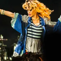 Rihanna na koncercie w Polsce, w bluzie z kolorowymi pasami, śpiewa z mikrofonem w dłoni.